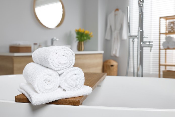 A stack of white towels on a wooden bath tray with a modern bathroom in the background, featuring a mirror, plants, and a robe.