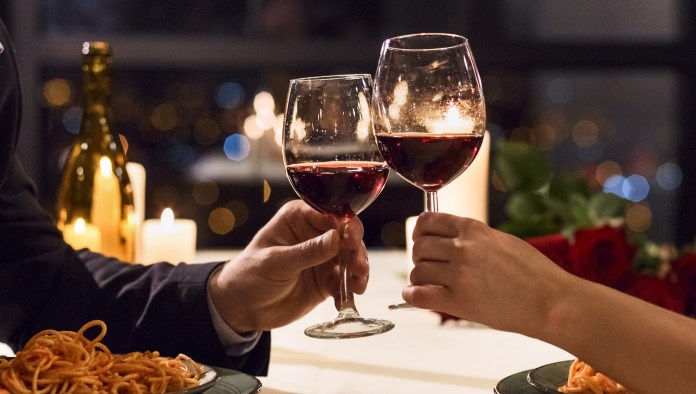 A romantic dinner scene featuring two people toasting with glasses of red wine, with a plate of spaghetti and candles in the background.