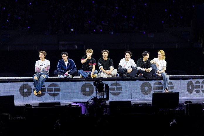 A group of eight male performers sitting together on a stage during a concert, wearing a mix of casual clothing. The background shows blurred crowd lights, contributing to the concert atmosphere.