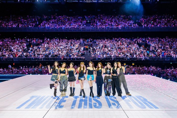 A South Korean girl group, TWICE, poses on stage in front of a cheering crowd during a concert, with the text 'TWICE THIS IS FOR' visible on the stage.