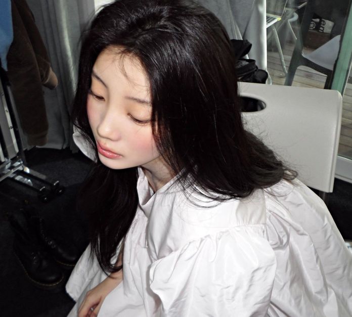 A young woman with long black hair sitting in a chair, wearing a white oversized blouse, looking down thoughtfully.