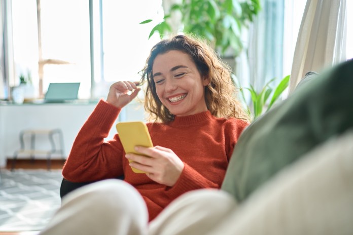A woman with curly hair smiles while sitting on a couch, holding a yellow smartphone in her hand, enjoying a moment in a bright and cozy indoor space.