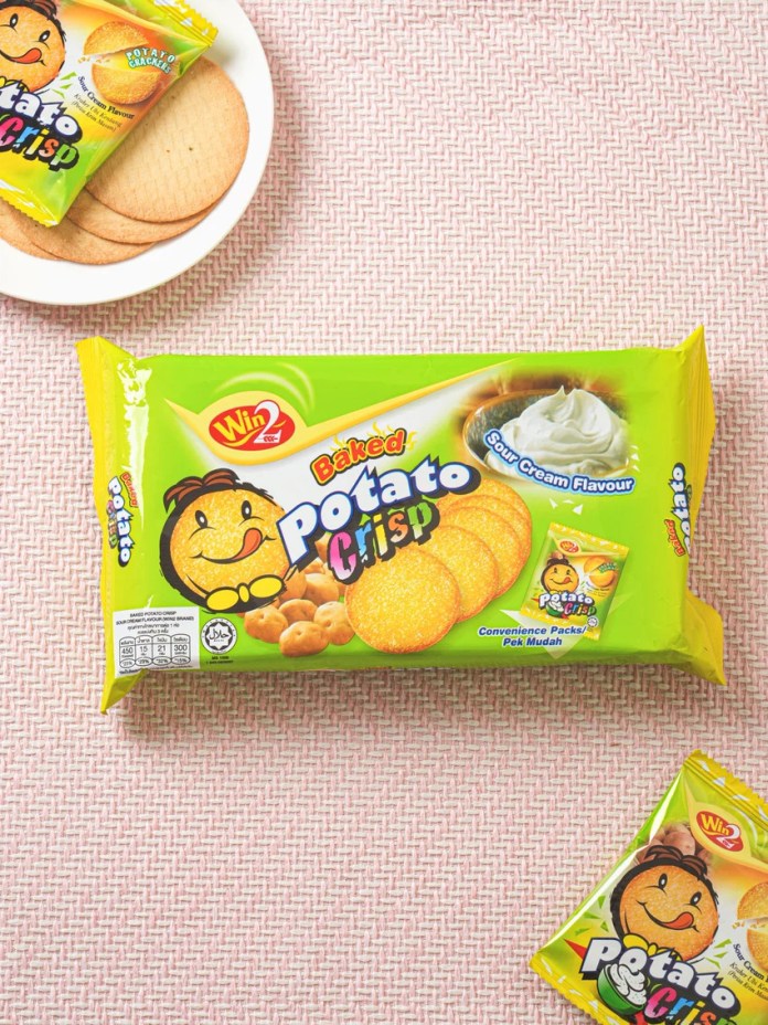 A package of baked potato crisps featuring a smiling cartoon character, with a bowl of round crackers shown on a pink textured surface.