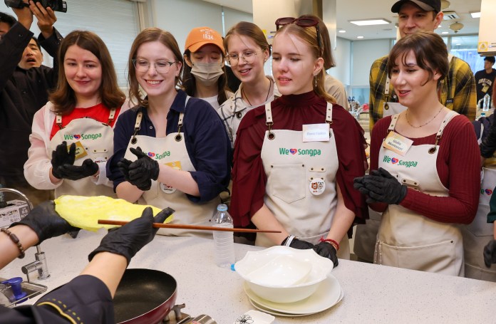 A group of five women in aprons watches intently as a chef demonstrates how to cook an omelet in a kitchen setting. They are engaged and smiling, with some wearing gloves.