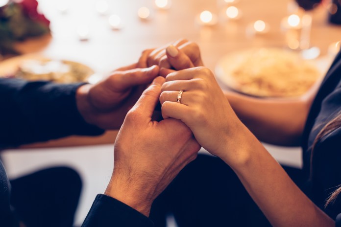 Close-up of two hands holding each other, one with a ring, set at a romantic dinner table with candles and food in the background.