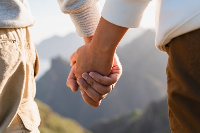 Two people holding hands while standing on a mountainside, with a scenic view of mountains in the background.