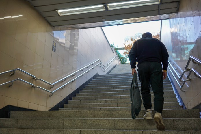 A person ascending a staircase in an underground station, holding a bag and wearing a dark hoodie.