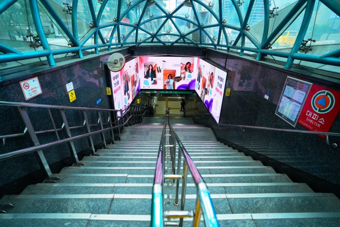 A view of a subway stairway leading down, featuring illuminated advertising displays on the walls and a glass canopy overhead.