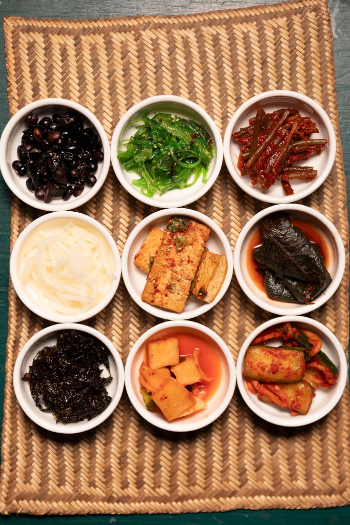 An assortment of side dishes served in small white bowls on a woven mat. The dishes include black beans, seasoned seaweed, pickled radish, tofu, spicy cucumbers, and fermented vegetables, creating a colorful and diverse spread.