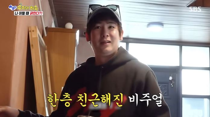 Nichkhun wearing a black hoodie and sunglasses is standing indoors, smiling at the camera. The background reveals a room with wooden paneling and a door. Text in yellow reads '친근해진 비주얼'.