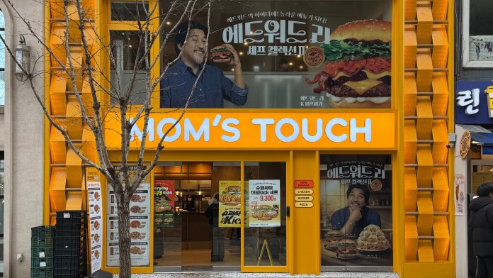 Exterior view of a bright orange restaurant called 'MOM'S TOUCH' featuring advertisements for burgers and fried chicken, with a smiling man holding a burger. The storefront has large windows and a tree in front.