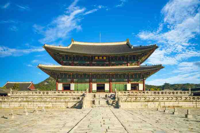 A traditional East Asian building with a multi-tiered roof, set against a blue sky with clouds, surrounded by stone pathways and greenery.