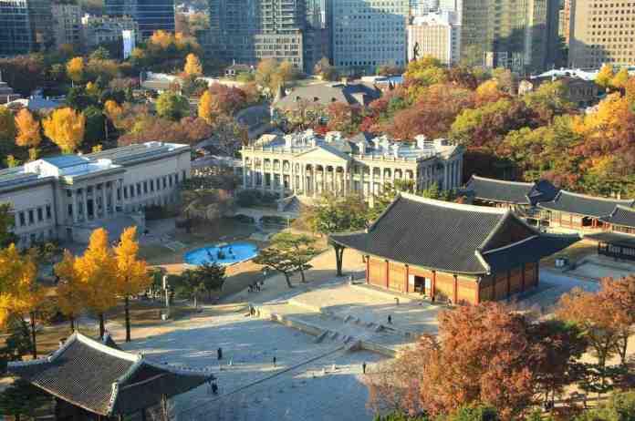 Aerial view of a park in autumn featuring colorful trees, a historical building with traditional architecture, and a modern city skyline in the background.