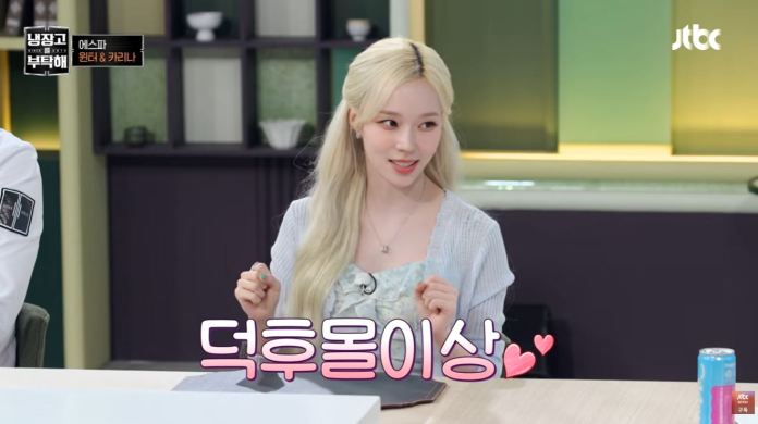 Winter with long blonde hair is sitting at a table, smiling and gesturing playfully. She is wearing a light blue top and has a friendly expression. The background features decorative elements, and a subtitle in pink text is visible in front of her.