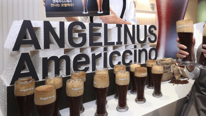 A display featuring various glasses of Americcino coffee drinks, with a prominent sign reading 'ANGELINUS Americcino' in bold letters. The background shows individuals holding drinks and coffee beans in burlap sacks.