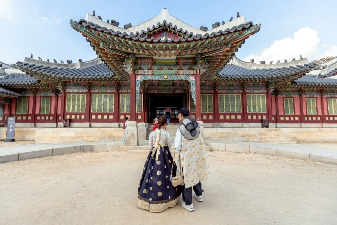 Two individuals wearing traditional Korean hanbok walking towards a historic building with ornate architecture in a cultural site.