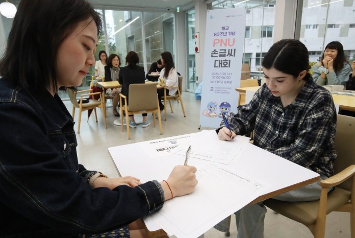 Two young women are focused on writing during an event in a bright, modern indoor space, with others seated in the background.