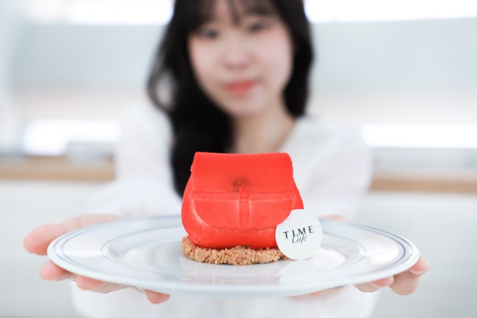 A person holding a plate with a red handbag-shaped dessert, featuring a round tag that reads 'TIME Cafe'. The background is softly blurred, emphasizing the dessert.