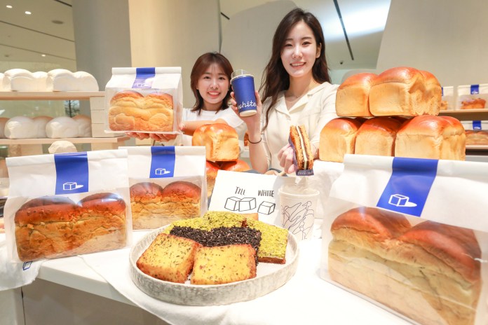 Two women smiling while showcasing various loaves of bread and baked goods at a bakery display.