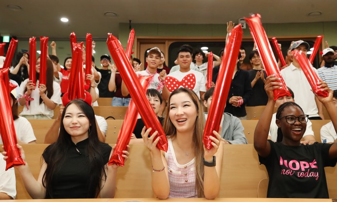 A diverse group of young people in a cheering section, holding red inflatable noisemakers while smiling and enjoying an event.