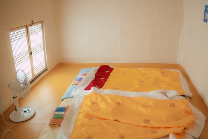 A bright, small room with two neatly arranged beds covered in colorful bedding. A fan is placed next to the beds, and a window with wooden slats allows soft natural light to enter.