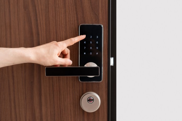 A close-up view of a hand pressing a number on a digital touchscreen keypad on a modern door lock, with a wooden door in the background.