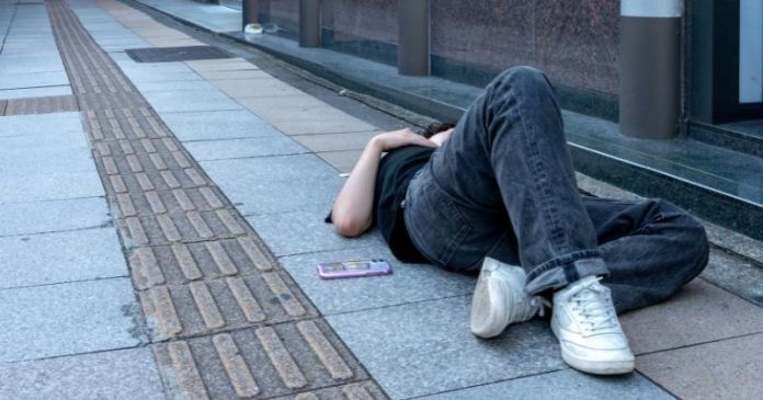 A person lying on the sidewalk with their back to the ground, wearing black jeans and white sneakers, with a phone placed nearby.
