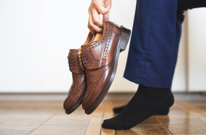 A person holds a pair of brown dress shoes while preparing to put them on, wearing dark socks and formal pants.