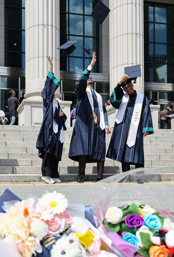 Three graduates in caps and gowns joyfully tossing their hats into the air in front of a university building, with a bouquet of flowers in the foreground.