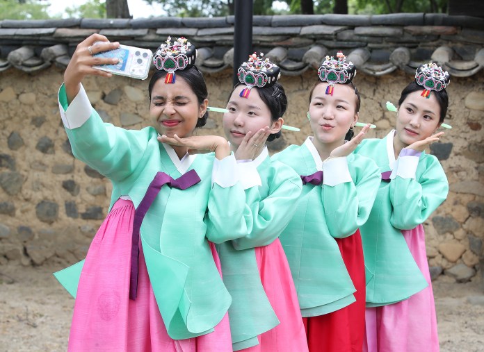 Four young women in traditional Korean hanbok outfits posing for a selfie, with smiles and playful expressions, against a rustic stone wall background.