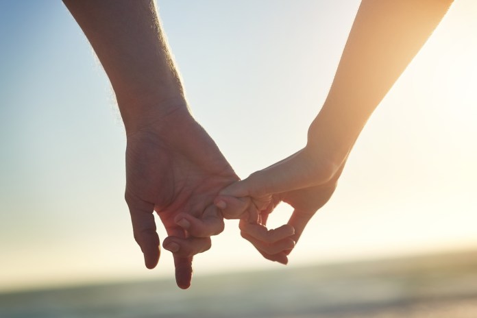 Close-up of two people holding hands against a sunset backdrop at the beach.