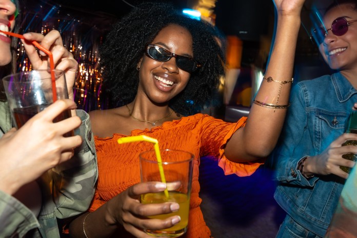 A group of friends enjoying a night out at a party, with one woman in an orange top smiling and raising her hand while holding a drink, surrounded by others also holding beverages.