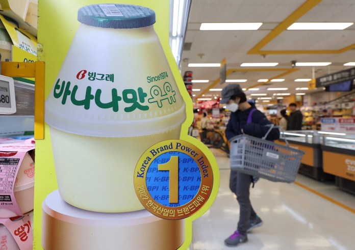 A bottle of banana-flavored milk in a grocery store, with a label indicating it is produced by Binggrae since 1974. A shopper in the background is carrying a basket.