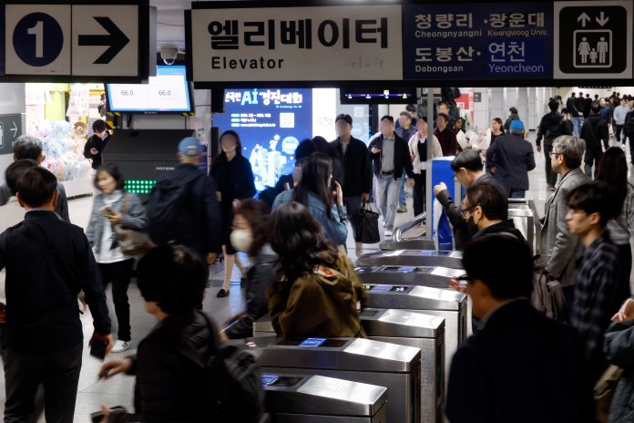 A busy subway station entrance with people passing through, featuring an 'Elevator' sign in Korean and English. Automated ticket barriers are in view, and digital displays show information. The crowd consists of various individuals, some using smartphones.