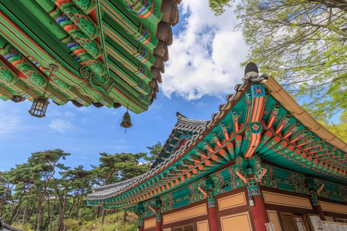 Colorful traditional Asian architecture featuring intricate wooden beams and roofs, with blue skies and trees in the background.