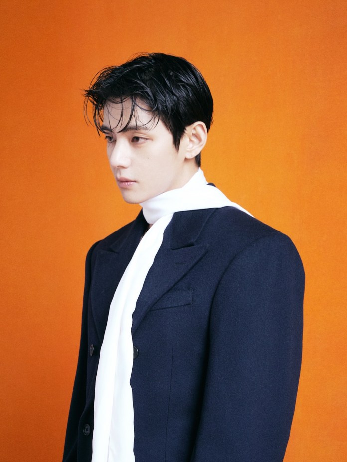 BTS V with dark hair wearing a navy coat and a white scarf, standing against an orange background.
