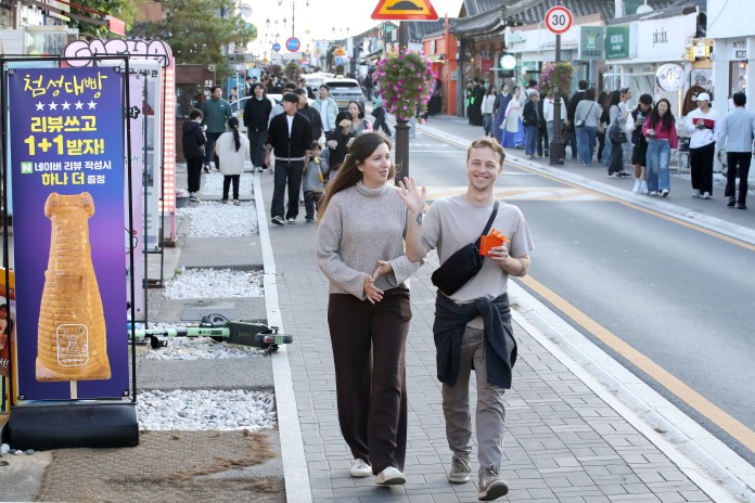 A couple walking hand-in-hand along a busy street, smiling and waving. They are surrounded by shops and people enjoying the area. A colorful sign advertising a food item is visible in the foreground.