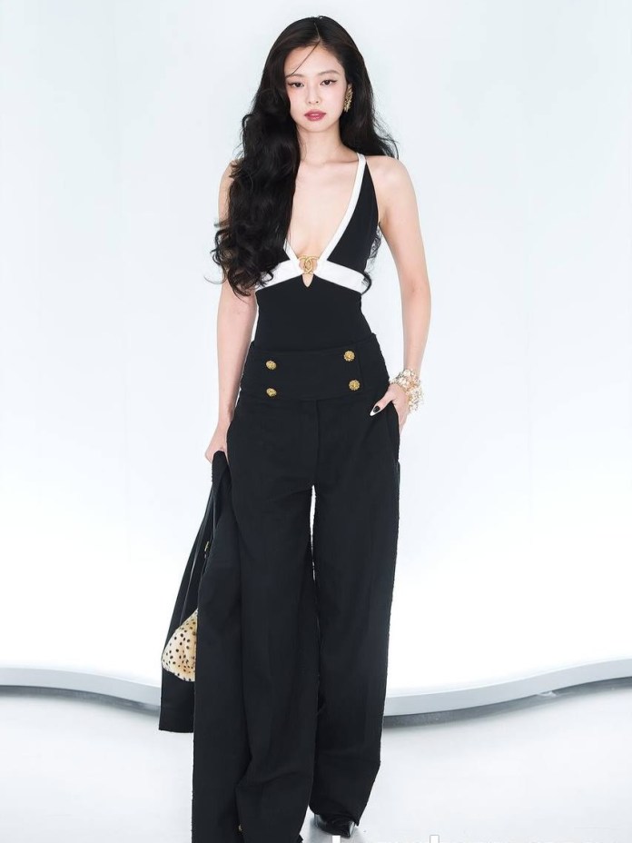Jennie stands confidently in a stylish black outfit featuring a deep V-neck and wide-leg trousers, accompanied by a handbag. The background is a soft, light color.