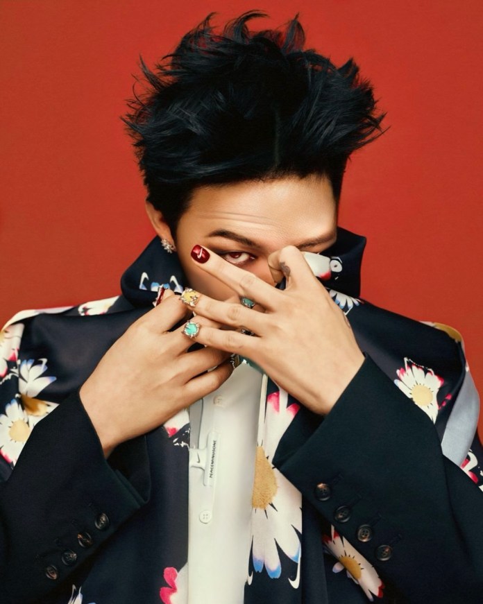 G-Dragon with stylish, spiky hair is partially obscuring their face with their hands, showcasing colorful nail art and rings. They are wearing a floral-patterned jacket against a red background.