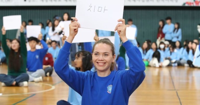 A smiling woman in a blue sweatshirt holds up a sign while sitting among a group of people in a gymnasium setting.