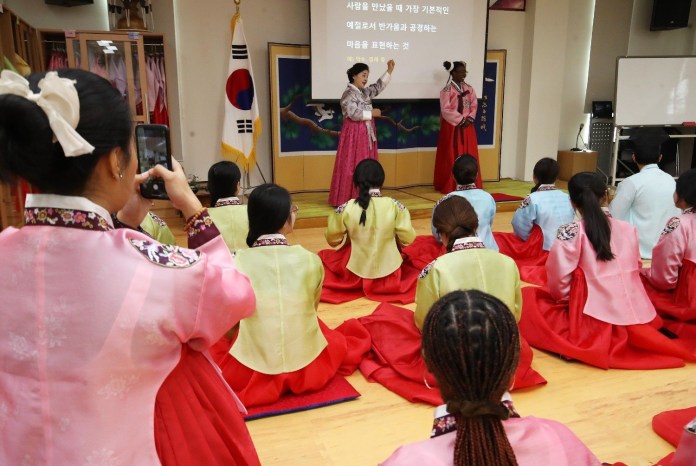 A group of people wearing traditional Korean hanbok sit on the floor, watching two individuals performing on stage. One performer gestures animatedly while the other stands beside her. The setting features a South Korean flag and a colorful backdrop.