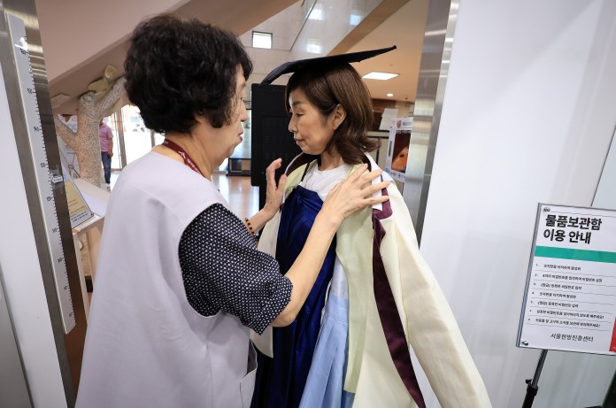 A woman in a graduation gown and cap is being assisted by another woman in a professional setting.