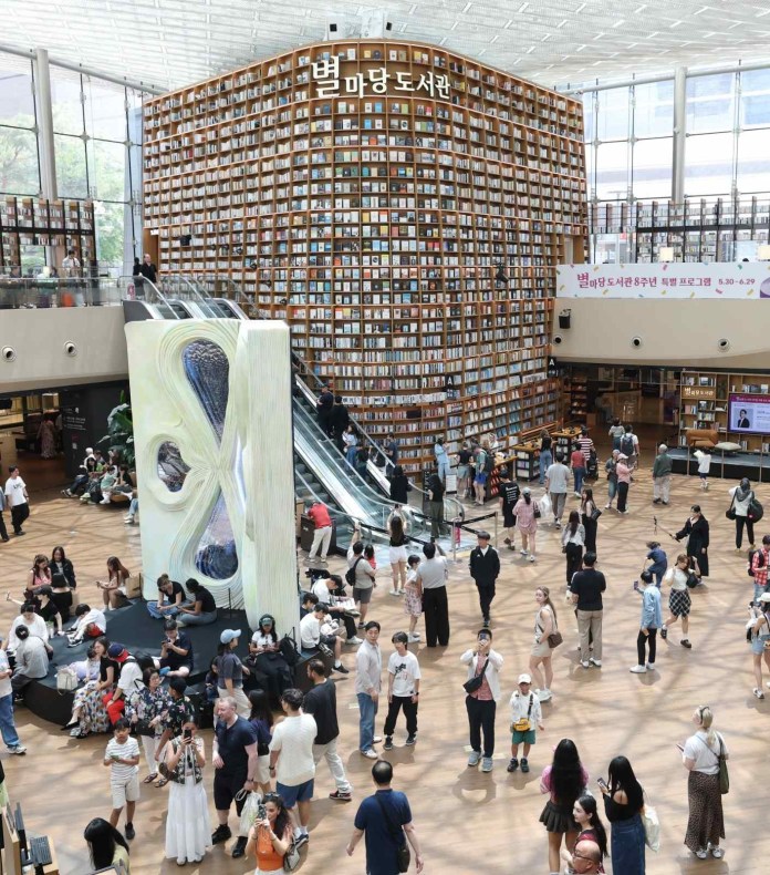A spacious library interior with a towering wall of books, many people walking around and reading, featuring escalators and modern design elements.