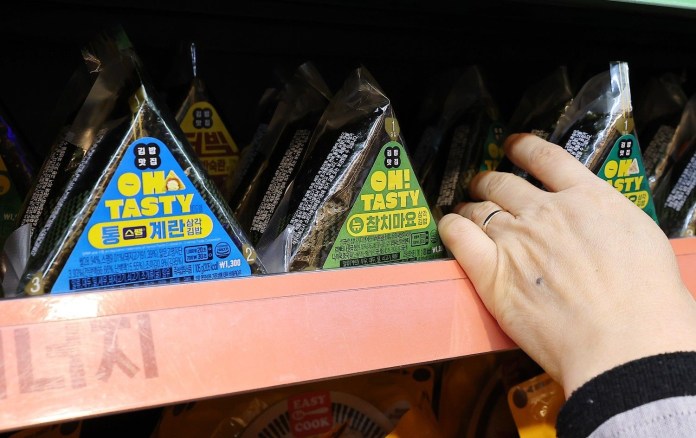 A hand reaching for a triangular package of 'Oh Tasty' seaweed snacks on a store shelf, with various flavors visible.