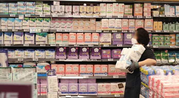 A person wearing a mask and gloves, holding several packages, stands in front of a shelf filled with various brands of paper products, organized by color and type.