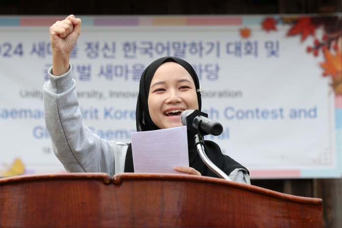A young girl wearing a hijab stands behind a podium, raising her fist in enthusiasm while speaking into a microphone. She appears confident and engaged, with a backdrop featuring a banner for a Korean language contest and gathering.