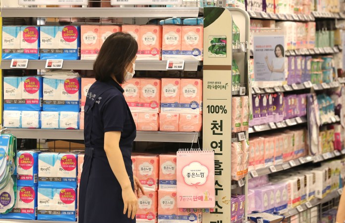 A shopper wearing a mask stands in an aisle of a supermarket, examining shelves filled with various brands of sanitary products, including packs in blue and pink packaging.