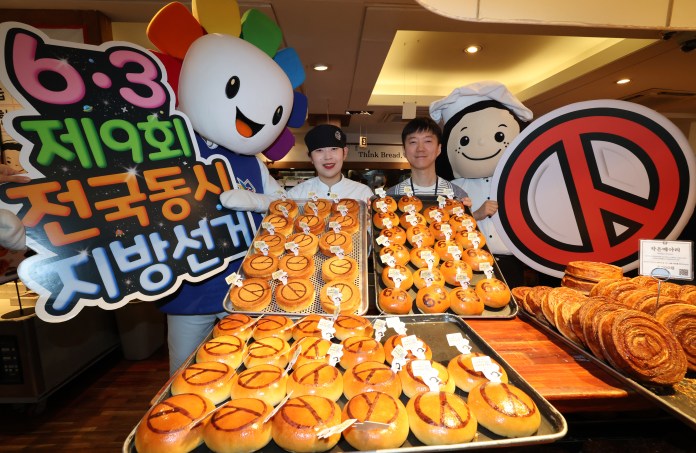 A cheerful mascot holds a sign for a national event while two bakers pose with trays of pastries decorated with a symbol. The background shows a bakery setting with various baked goods.