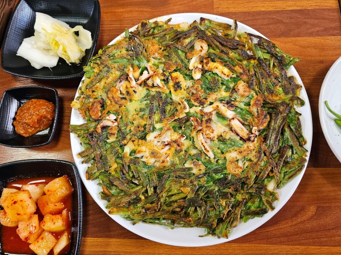 A plate of green vegetable pancakes, garnished with pieces of seafood, served alongside small black bowls of dipping sauce and pickled vegetables, on a wooden table.