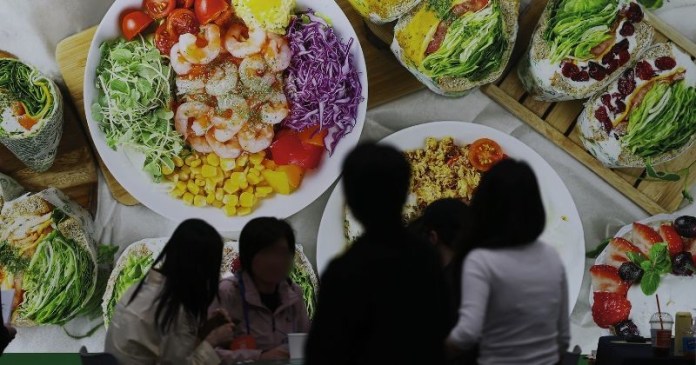 A vibrant display of fresh food including shrimp, vegetables, and fruit, with people silhouetted in the foreground.
